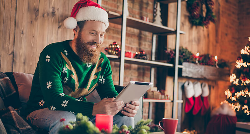 Guy sitting on comfy chair browsing his tablet and wearing a Santa hat at home.