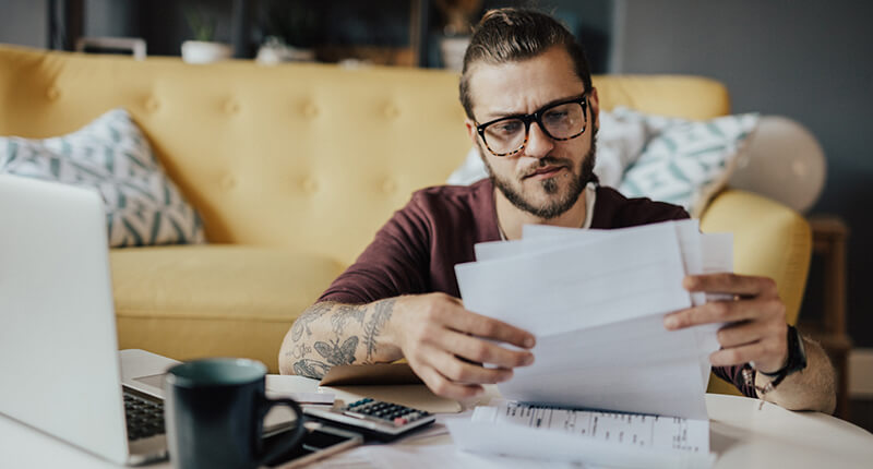 Millennial man studies bills at his desk with computer open, wondering how to pay down debt.